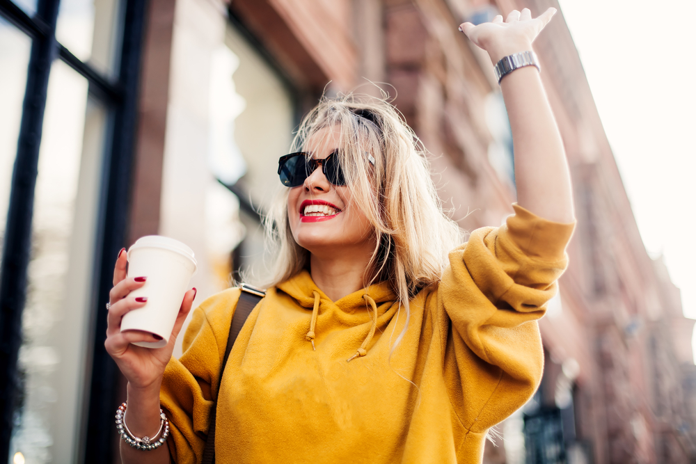 Excited blonde woman holding coffee while walking and raising hand